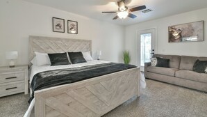 A bedroom with a bed, a ceiling fan, a nightstand, a bedside table, a couch, and a door.