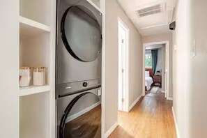 Un cuarto de lavandería con lavadora y secadora, un pasillo que conduce a un dormitorio, y una puerta a otra habitación.