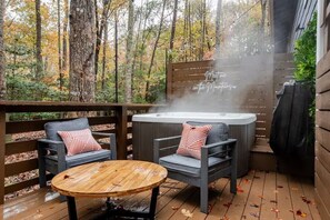 A hot tub with steam, two gray chairs with red and white pillows, and a wooden table on a deck with a wooden fence and trees in the background.