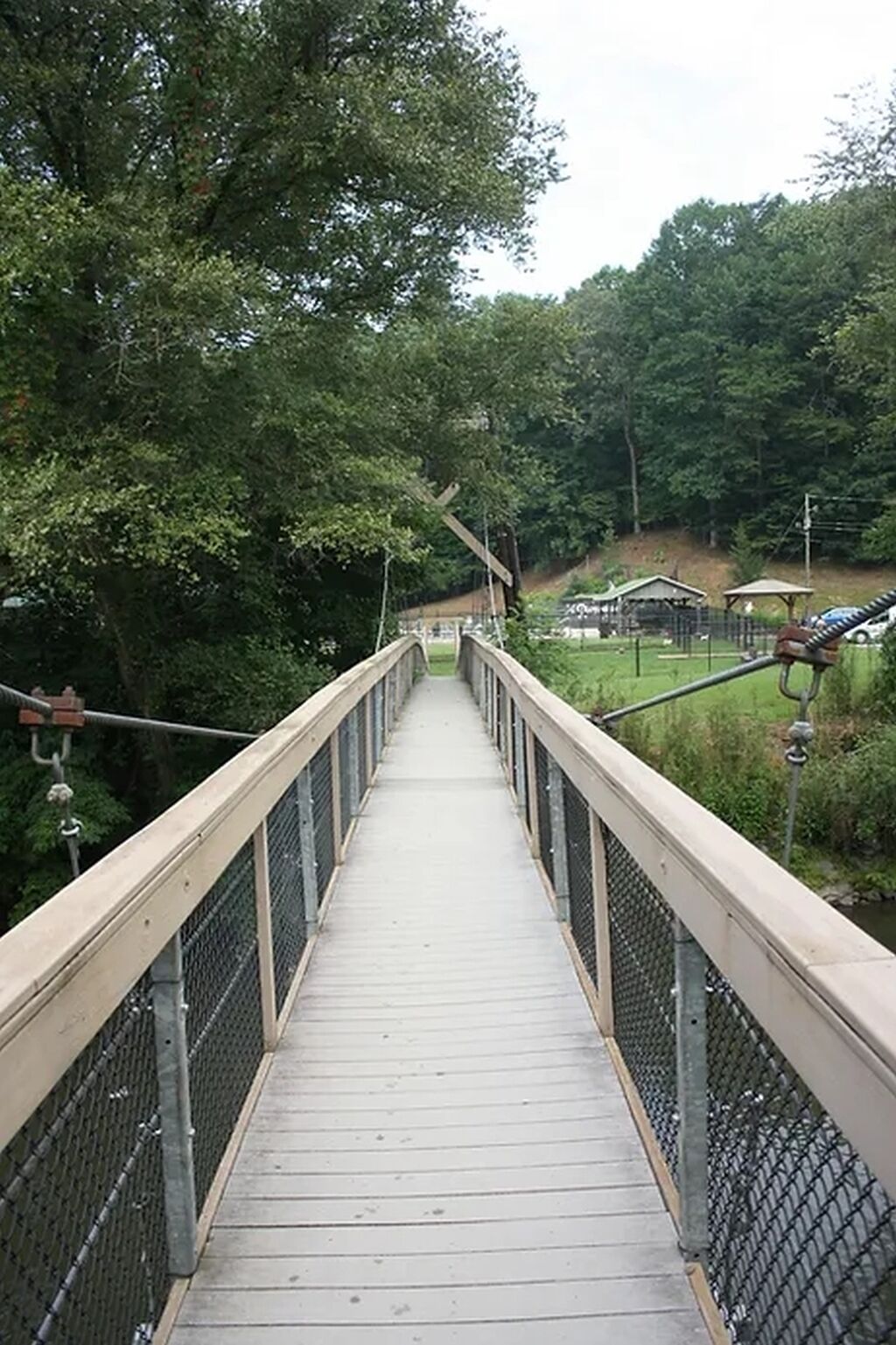 A wooden pedestrian bridge with metal railings, surrounded by trees and a grassy area.