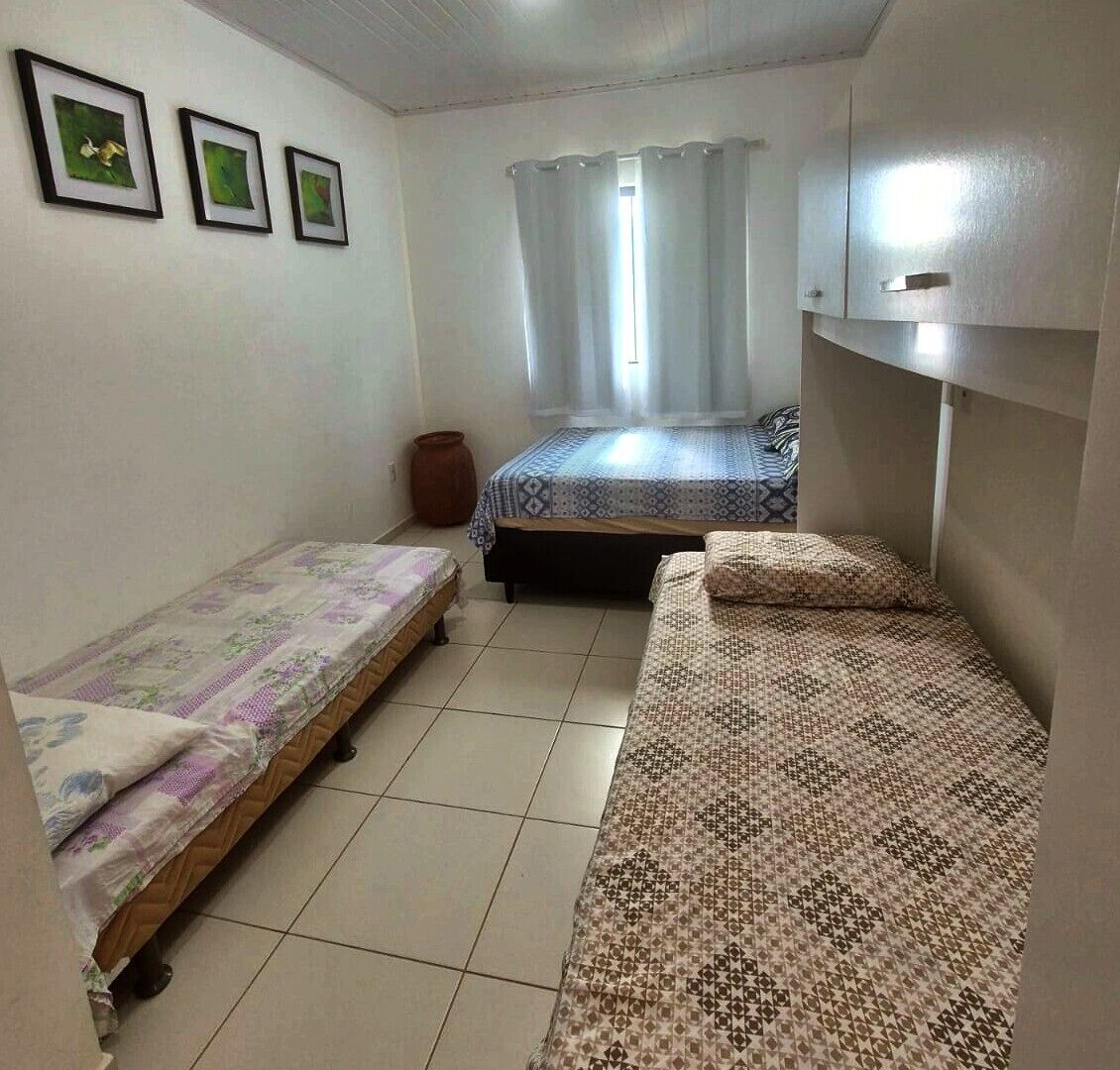 Quarto pequeno e limpo com duas camas de solteiro, uma janela com cortinas e uma prateleira na parede.