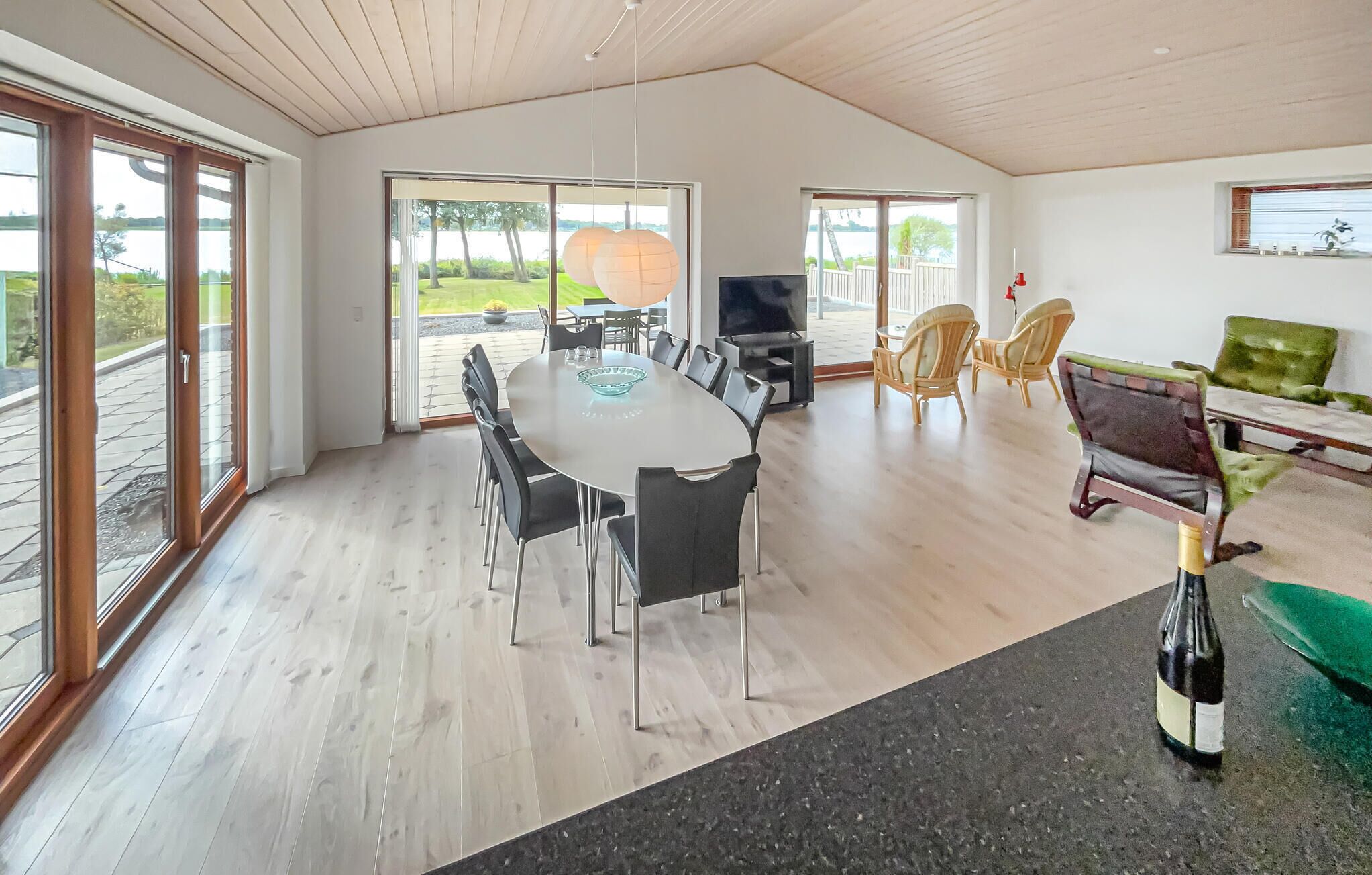 A spacious dining area with a large table and chairs, a TV, and a view of a lake through large windows.