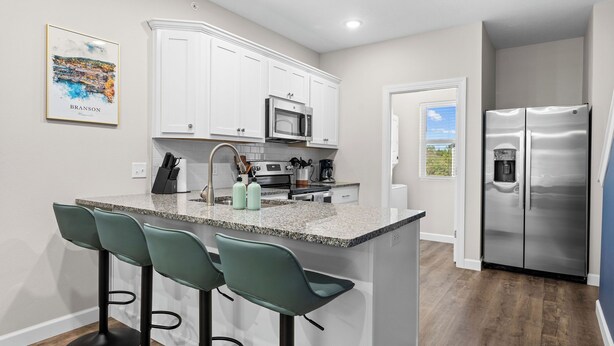 A modern kitchen with white cabinets, a granite countertop, and a stainless steel refrigerator. There are three bar stools and a kitchen island. - Perfect Location! Near SDC, Branson Strip & Lake (Branson)