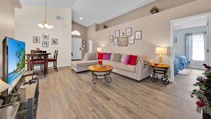 Ein Wohnzimmer mit Sofa, Couchtisch und Fernseher. An den Wänden hängen gerahmte Bilder, und es gibt einen Essbereich mit Stühlen und Tisch.