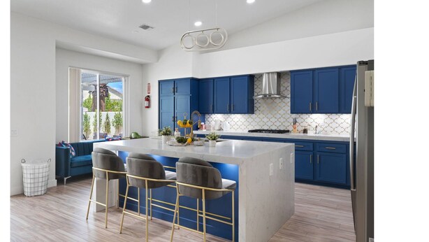 Een moderne keuken met blauwe kastjes, een centraal eiland met barstoelen en een groot raam.