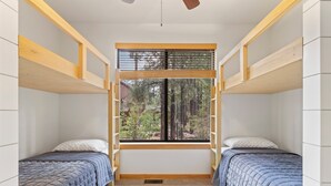 Une chambre avec deux lits superposés, une grande fenêtre avec des stores et une vue sur les arbres à l’extérieur.