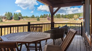 A wooden deck with a table and chairs, overlooking a grassy area with trees and buildings. - Alpenglow Aura in Angel Fire (Angel Fire)