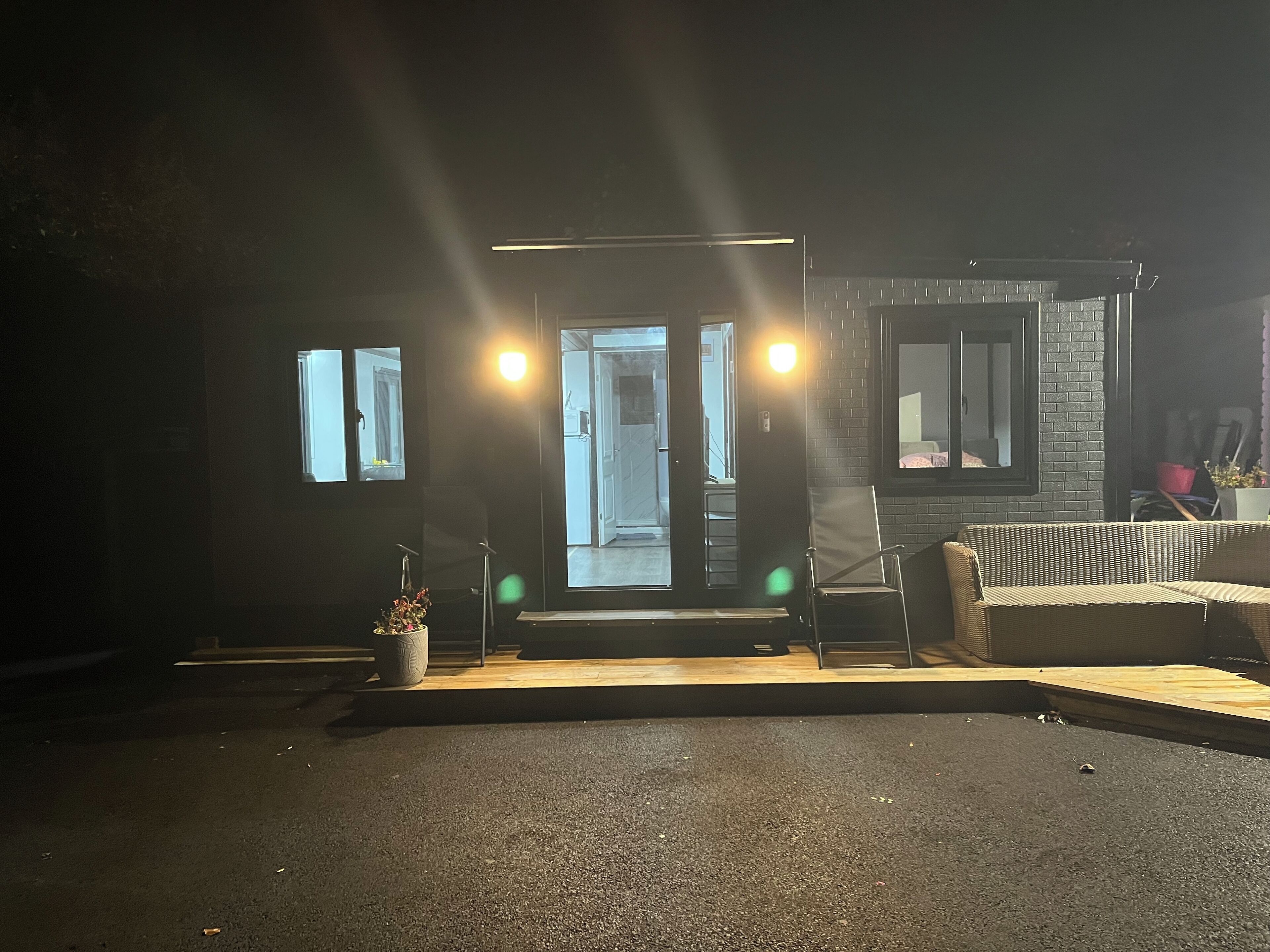 Un bâtiment à la façade sombre, doté de deux fenêtres illuminées et d’une porte donnant sur un espace intérieur.