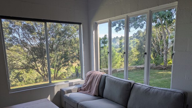 A room with a grey sofa, a small table, and large windows offering a view of a green landscape with trees. - Maleny-Montville Cottages #4 - 2 bed with ocean views (Balmoral Ridge)