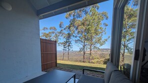 A room with a view of a grassy landscape and trees, a wooden fence, and a black table and grey sofa. - Maleny-Montville Cottages #4 - 2 bed with ocean views (Balmoral Ridge)