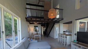 A spacious living area with a large sofa, a wooden coffee table, and a TV on a white stand. There is a staircase leading to an upper level with a balcony. - Maleny-Montville Cottages #4 - 2 bed with ocean views (Balmoral Ridge)