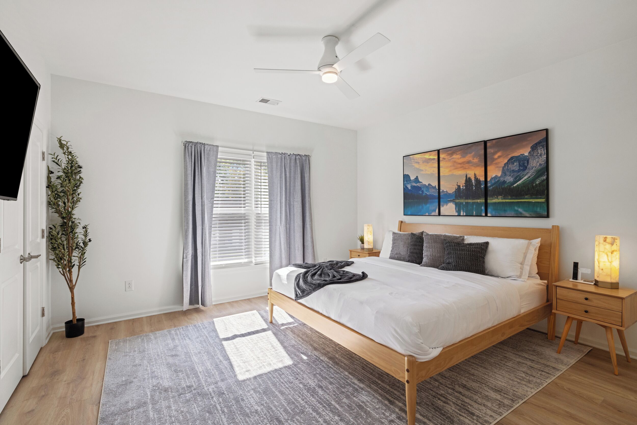 Ein Schlafzimmer mit einem hölzernen Bett, einem Deckenventilator, einem großen Fenster mit Vorhängen, einem Nachttisch mit Lampe und einem an der Wand hängenden Gemälde.