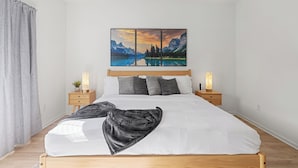 A bedroom with a wooden bed, white bedding, a gray throw blanket, bedside tables with lamps, and a wall-mounted painting of a mountain landscape. - The Stadium Getaway (Lexington)