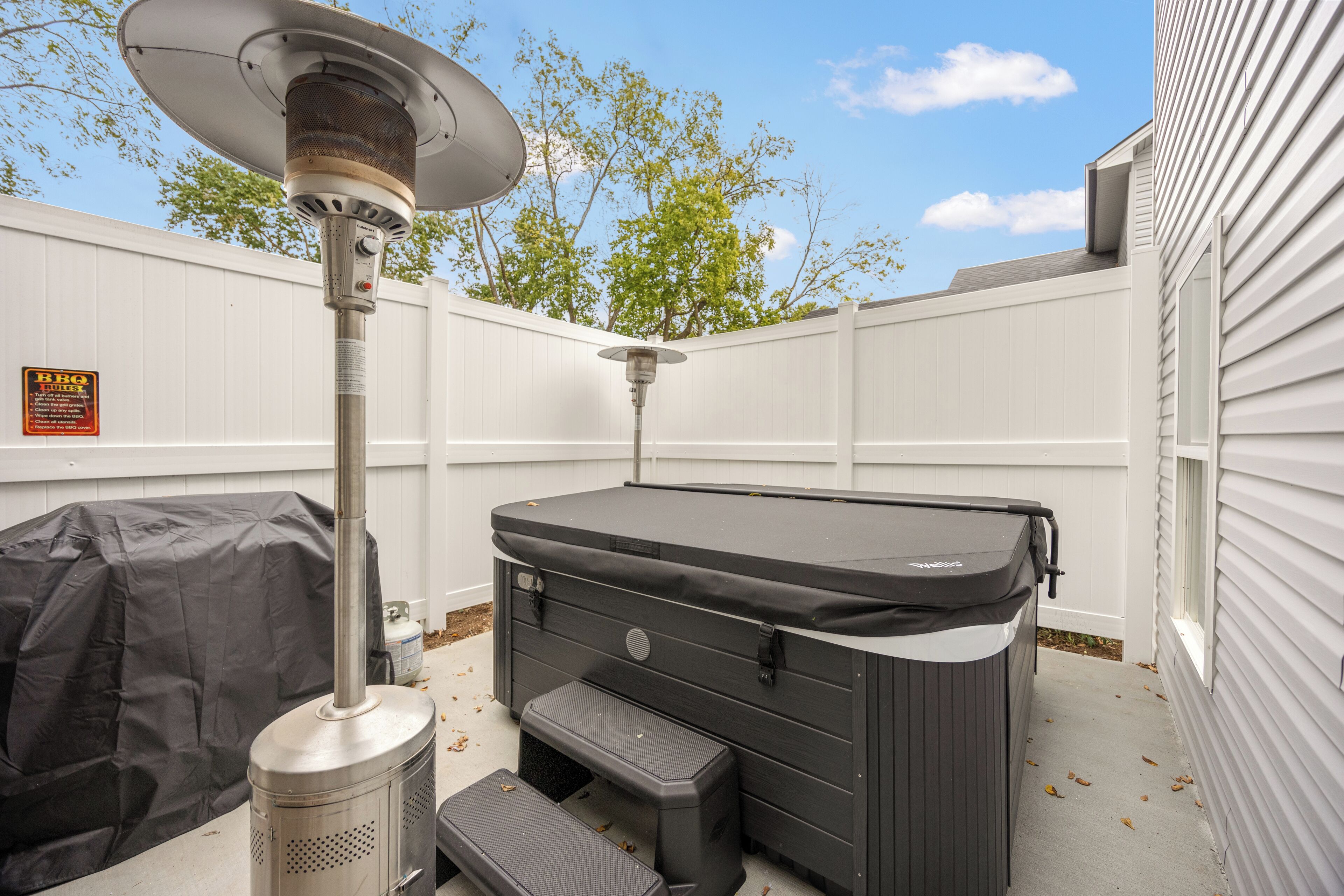 A portable hot tub with a cover and steps, set against a white fence and a house.