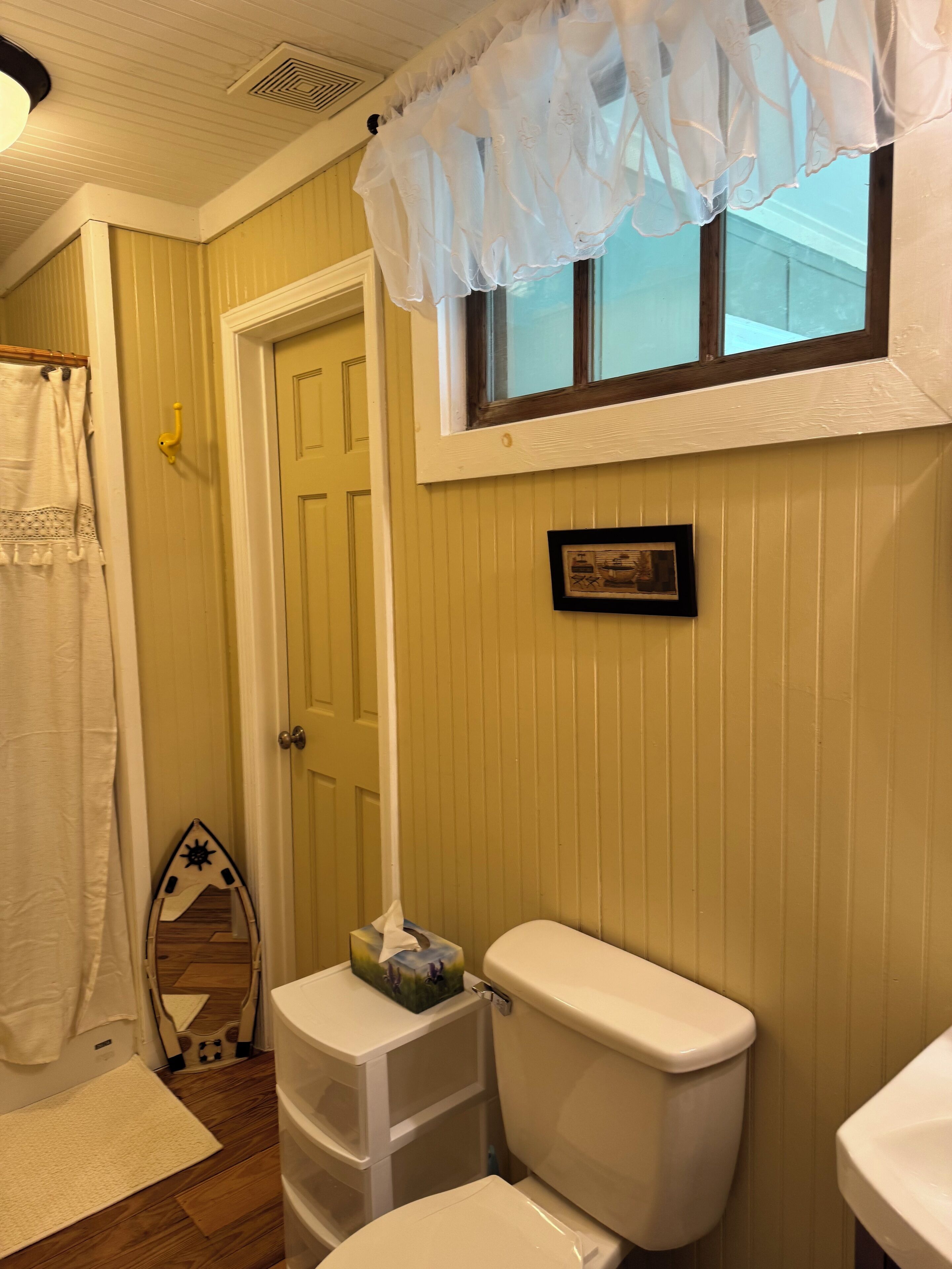 A bathroom with a toilet, a window with a white valance, a framed picture on the wall, and a towel hanging on a rack.