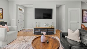 A living room with a grey sofa, a black leather couch, a wooden coffee table with a vase, a TV on the wall, and a sideboard with decorative items. - Spacious 3BR Retreat | Sleeps 8 | 15 Min Airport (Calgary)