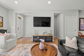 A living room with a grey sofa, a black leather couch, a wooden coffee table with a vase, a TV on the wall, and a sideboard with decorative items. - Spacious 3BR Retreat | Sleeps 8 | 15 Min Airport (Calgary)