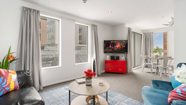 A modern living room with a flat-screen TV, a red cabinet, a dining area with a table and chairs, and large windows with curtains. - Perfect City Retreat 1 bdrm Prime Location (Christchurch)