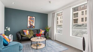 A modern living room with a blue sofa, a black leather couch, and a round coffee table. There is a large window with a view of buildings and a fan on the right. - Perfect City Retreat 1 bdrm Prime Location (Christchurch)