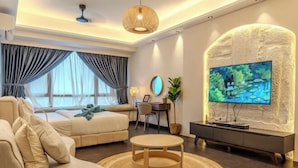 A modern living room with a sofa, a round coffee table, a TV mounted on a textured wall, a desk with a chair, and a large window with curtains. - R&F Princess Cove by Nest Home at Johor Bahru (Johor Bahru)