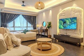 A modern living room with a sofa, a round coffee table, a TV mounted on a textured wall, a desk with a chair, and a large window with curtains. - R&F Princess Cove by Nest Home at Johor Bahru (Johor Bahru)