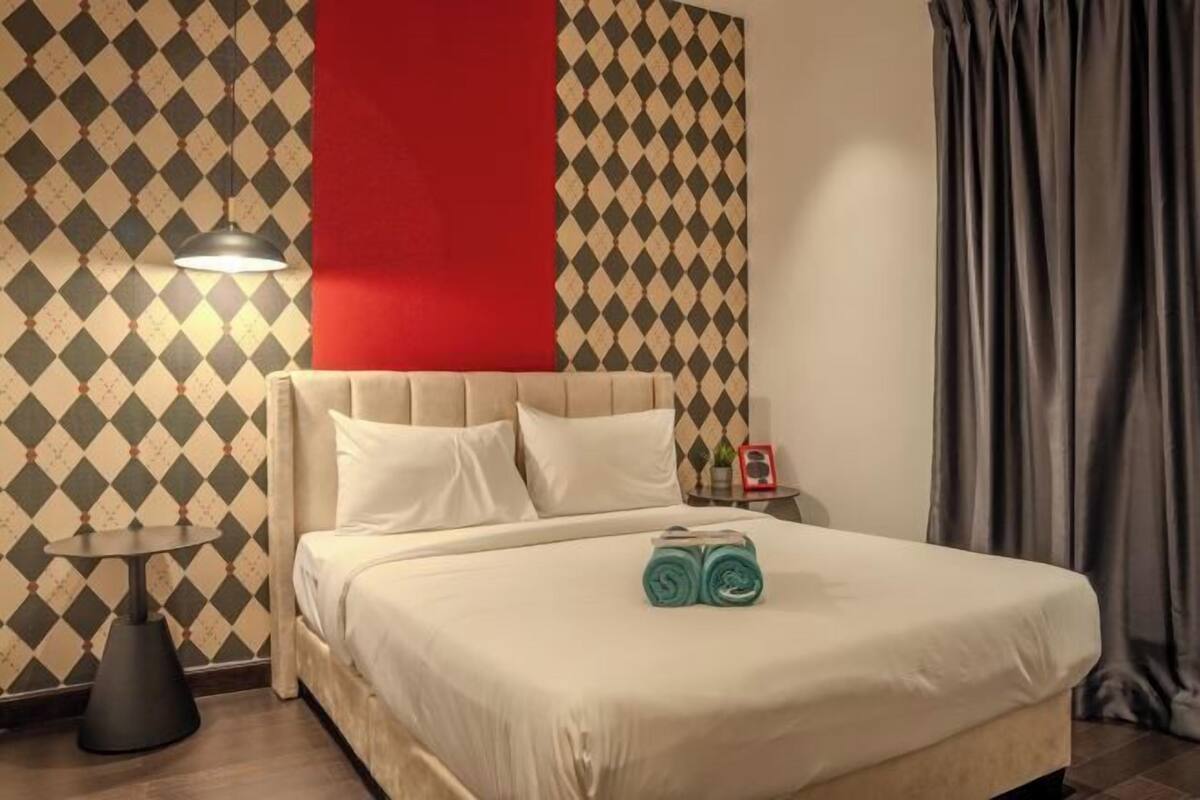A hotel room with a patterned wallpaper, a red accent wall, a bed with white bedding, a bedside table with a lamp, and a pair of rolled towels on the bed.