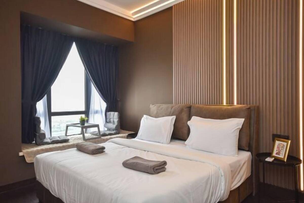 A hotel room with a large bed, a headboard, a small table with a plant, and a view of the outdoors through a window.