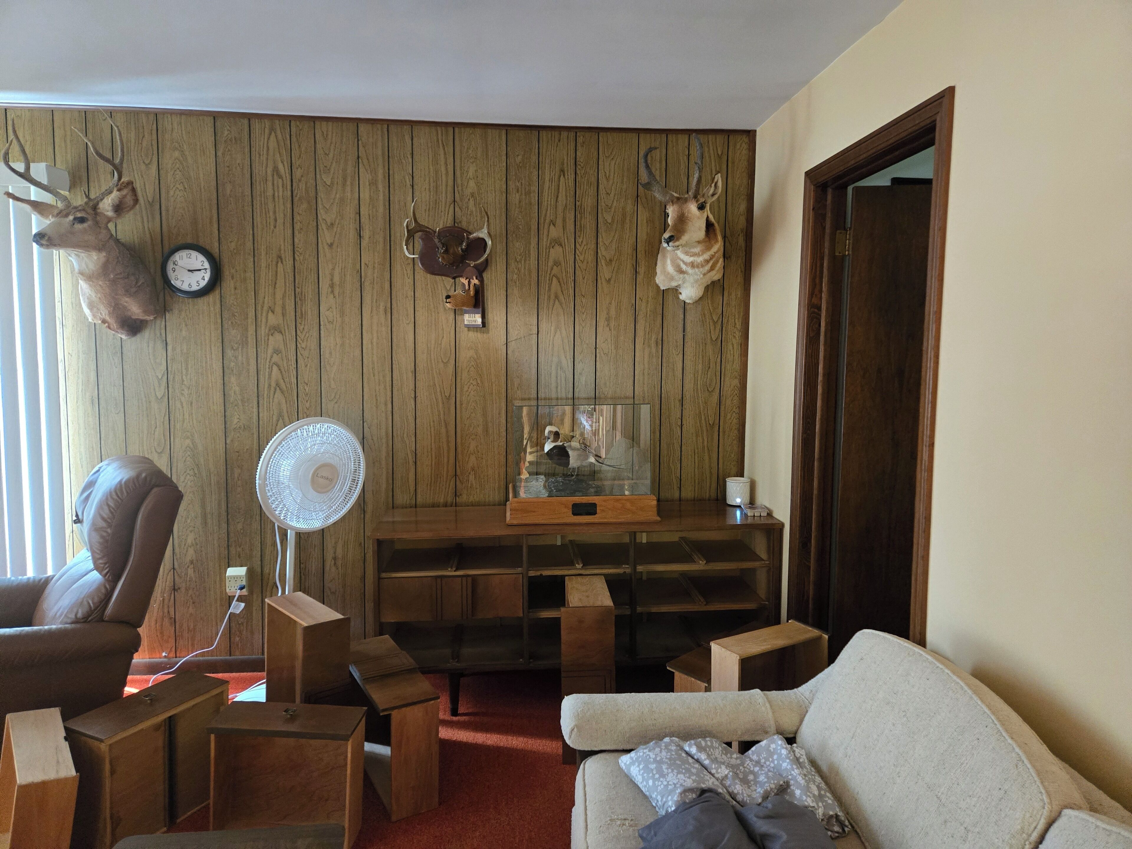 A living room with wooden panel walls, a clock, mounted deer heads, a television, and a fan.
