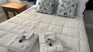 A neatly made bed with a quilted white duvet, two folded white towels with brown handles, and a wooden nightstand with a lamp.