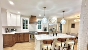 A modern kitchen with white cabinetry, a central island with bar stools, stainless steel appliances, and pendant lighting. - Renovated 3-bedroom house in Montgomery VT, close to Jay Peak (Montgomery Center)