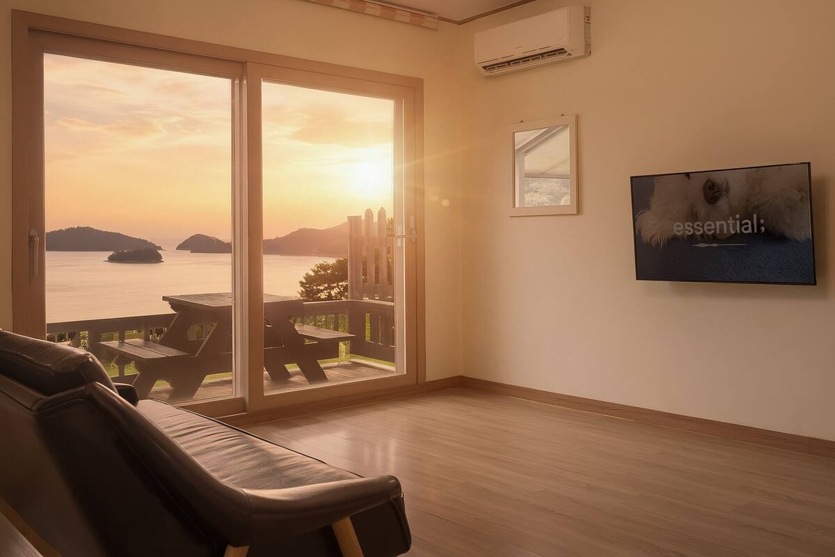 Ein Zimmer mit einer großen Schiebeglasdoor, die auf einen Balkon mit Blick auf das Meer und die Inseln führt. Es gibt ein Ledersofa und einen flachbildschirm-Fernseher an der Wand.