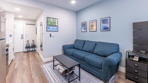 A modern living room with a blue sofa, a small table, and a coffee table. There are framed pictures on the wall and a coat rack near the door. - Slopeside Ski-In/Out Condo 112 | Snowshoe Village (Snowshoe)