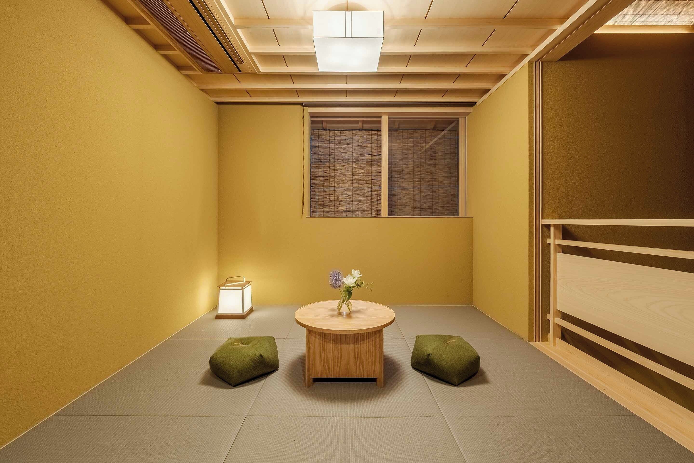 A room with tatami flooring, a round wooden table with a vase of flowers, two green cushions, a window with a view, and a ceiling light fixture.