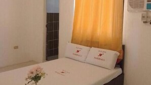 A hotel room with a bed, a window with yellow curtains, an air conditioner, and a small table with a vase of flowers. - WJV INN MARIBAGO (Lapu-Lapu)