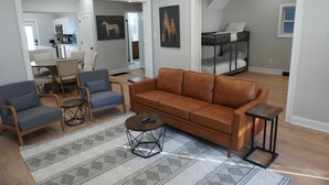 Un salon moderne avec un canapé en cuir marron, deux fauteuils bleus et une table basse. On aperçoit à l’arrière-plan un coin repas avec une table et des chaises.