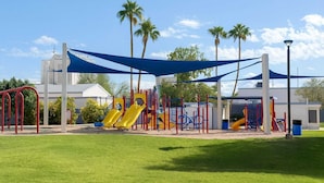 A playground with slides and climbing structures, surrounded by palm trees and a clear sky.