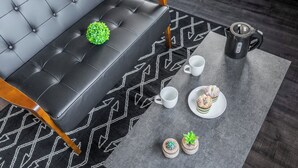 Ein modernes Wohnzimmer mit einem schwarzen Ledersofa, einem Teppich mit geometrischem Muster, einem Couchtisch, einer weißen Tasse, einem Teller mit Gebäck und zwei kleinen Topfpflanzen.