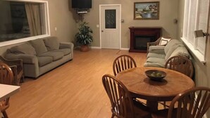 A living room with a sofa, a dining table with chairs, a television, a fireplace, and a window with blinds. - Creekside Chalet + Allstar Deluxe (Otego)