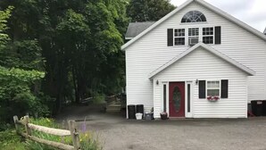 A white building with a red door, surrounded by greenery and a gravel driveway. - Creekside Chalet + Allstar Deluxe (Otego)