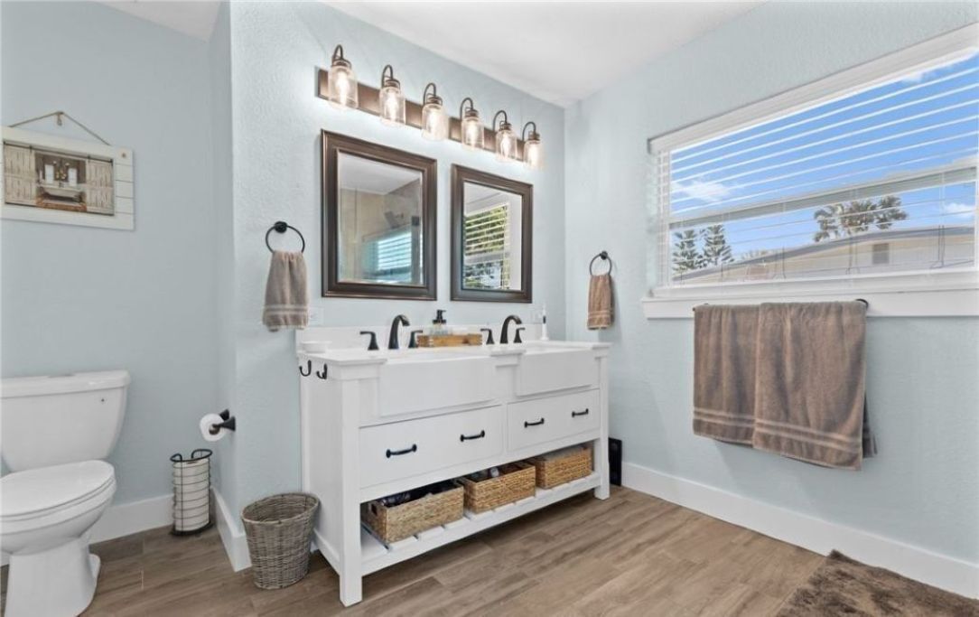 Une salle de bain avec un meuble-lavabo blanc, deux miroirs, une fenêtre avec des stores et un porte-serviettes.