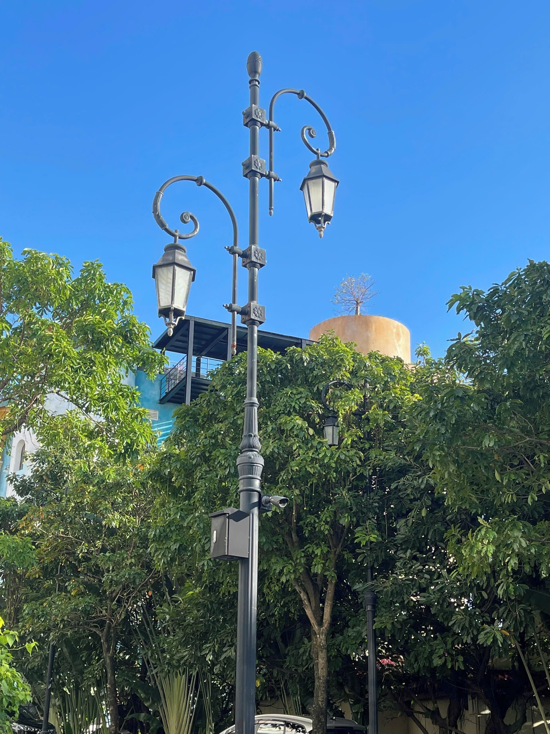 A street lamp with ornate design, surrounded by lush green trees and a building with a blue facade in the background.