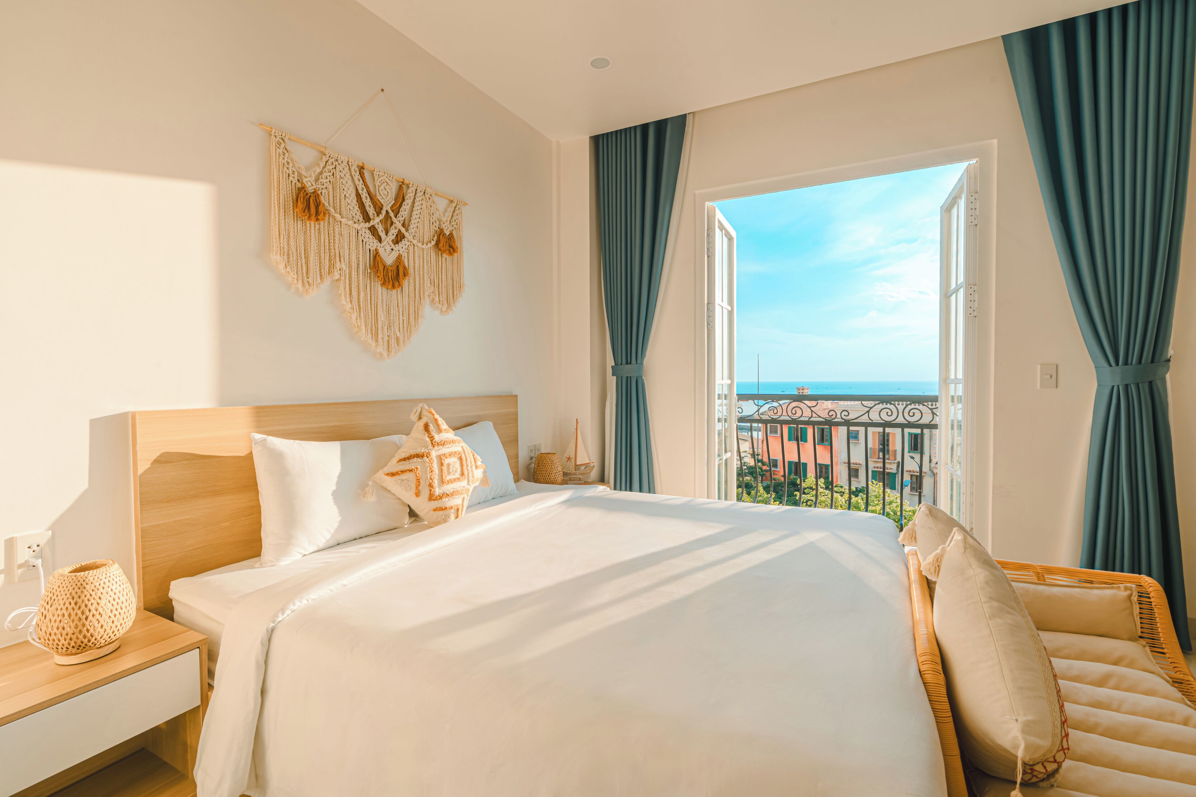 Quarto de hotel com uma cama grande, uma mesa de cabeceira, uma cadeira de vime e vista para a cidade através de uma janela aberta.