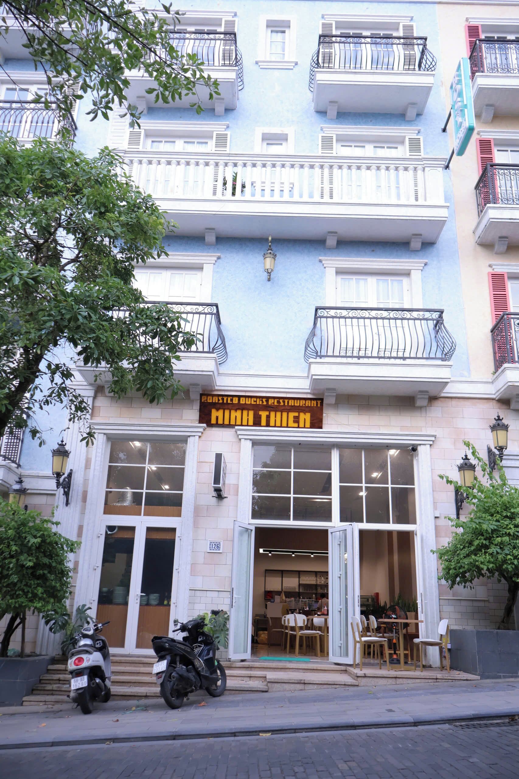 A restaurant with a sign reading "Roasted Duck's Restaurant" and "Minh Thiên". It has a white facade, multiple balconies, and a few parked scooters in front.