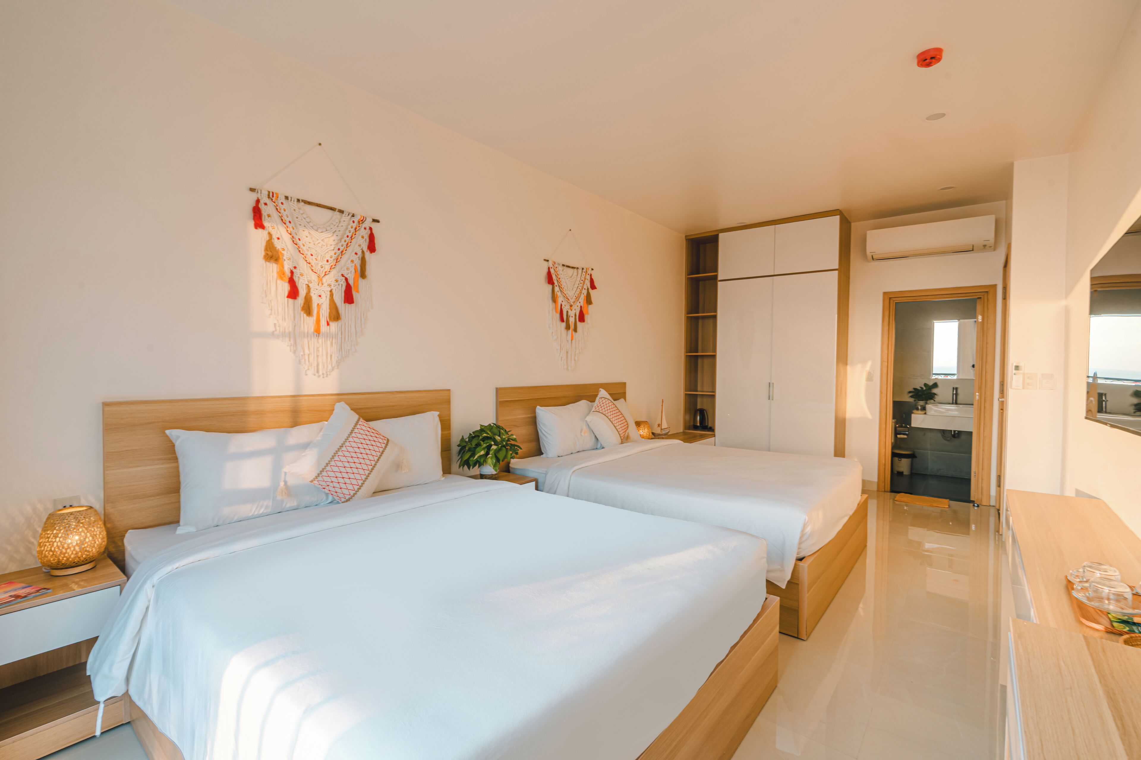 A modern hotel room with two beds, a wooden headboard, a bedside table with a lamp, and a small plant.