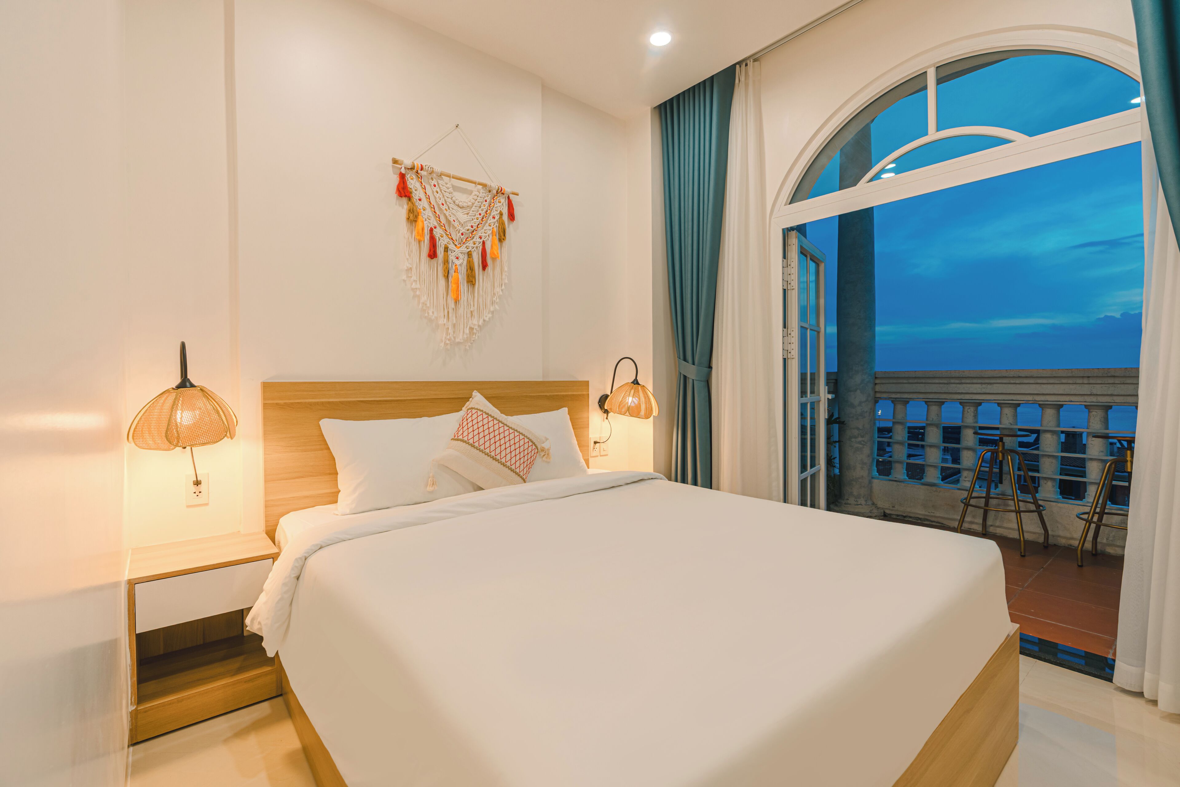 A hotel room with a large bed, bedside lamps, a decorative wall hanging, and a balcony with a view.