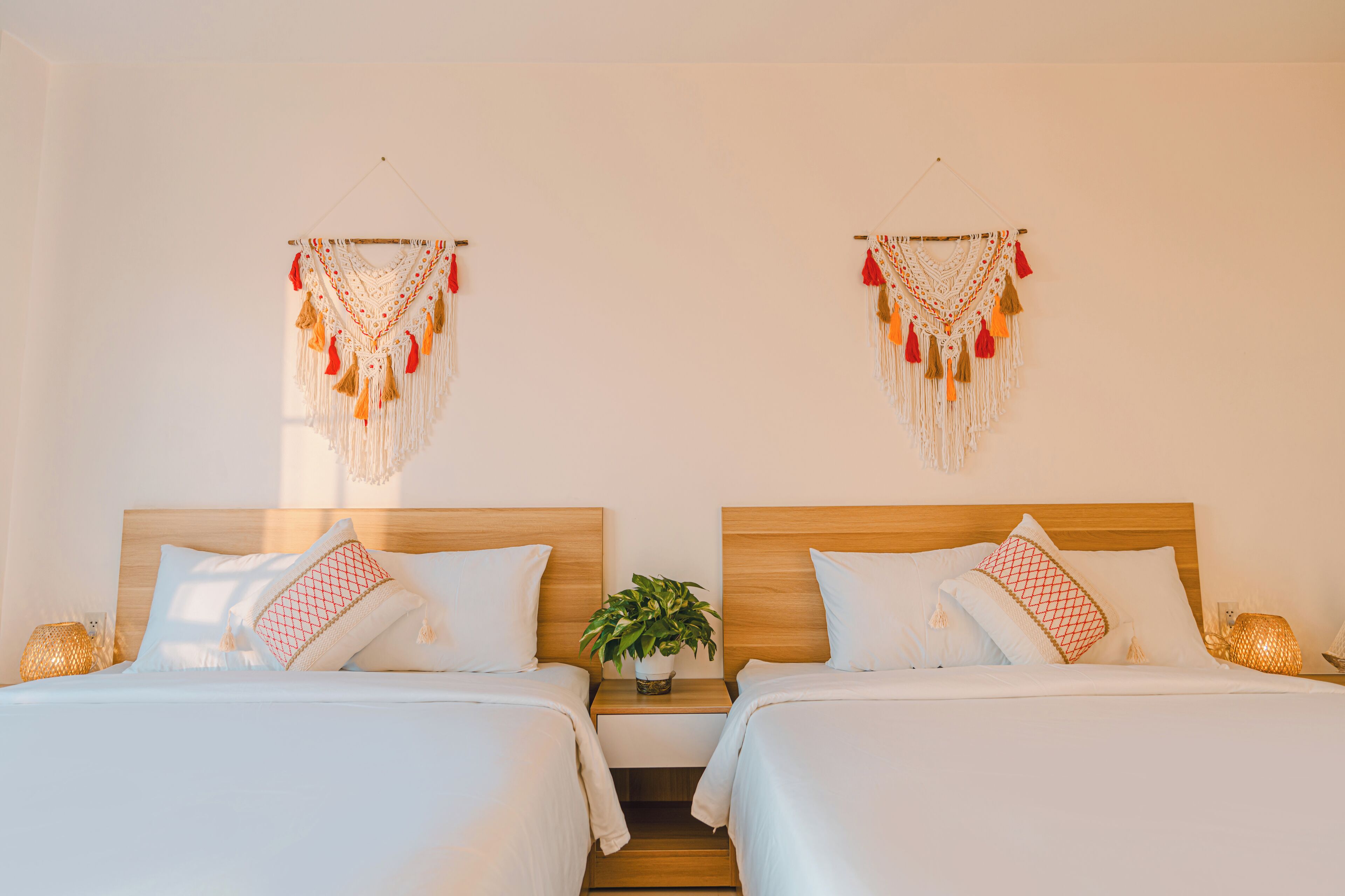 A hotel room with two beds, white bedding, a wooden headboard, a small bedside table with a plant, and wall decorations.