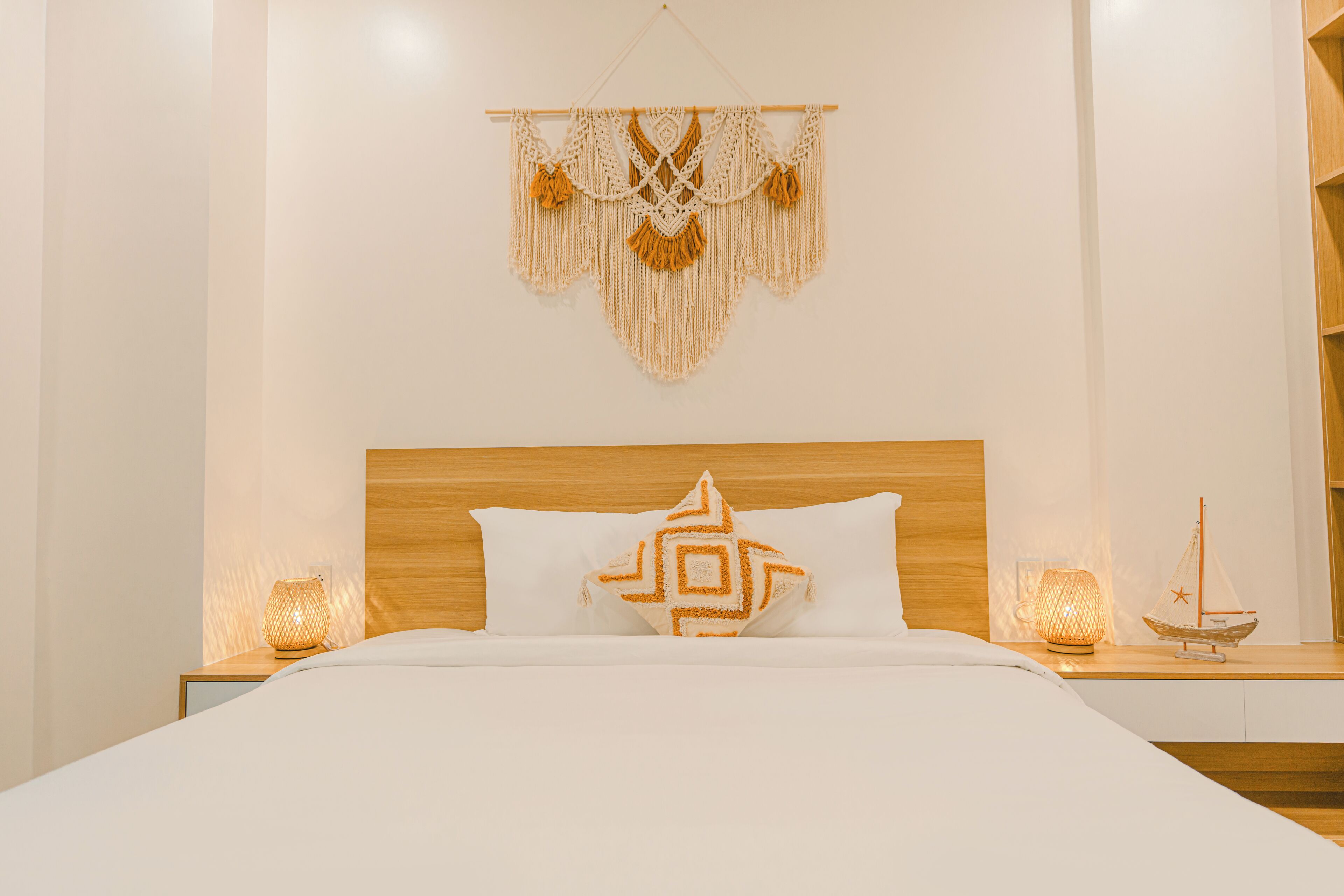 Una cama bien hecha con un cojín geométrico, un tapiz de macramé y dos lámparas de noche.
