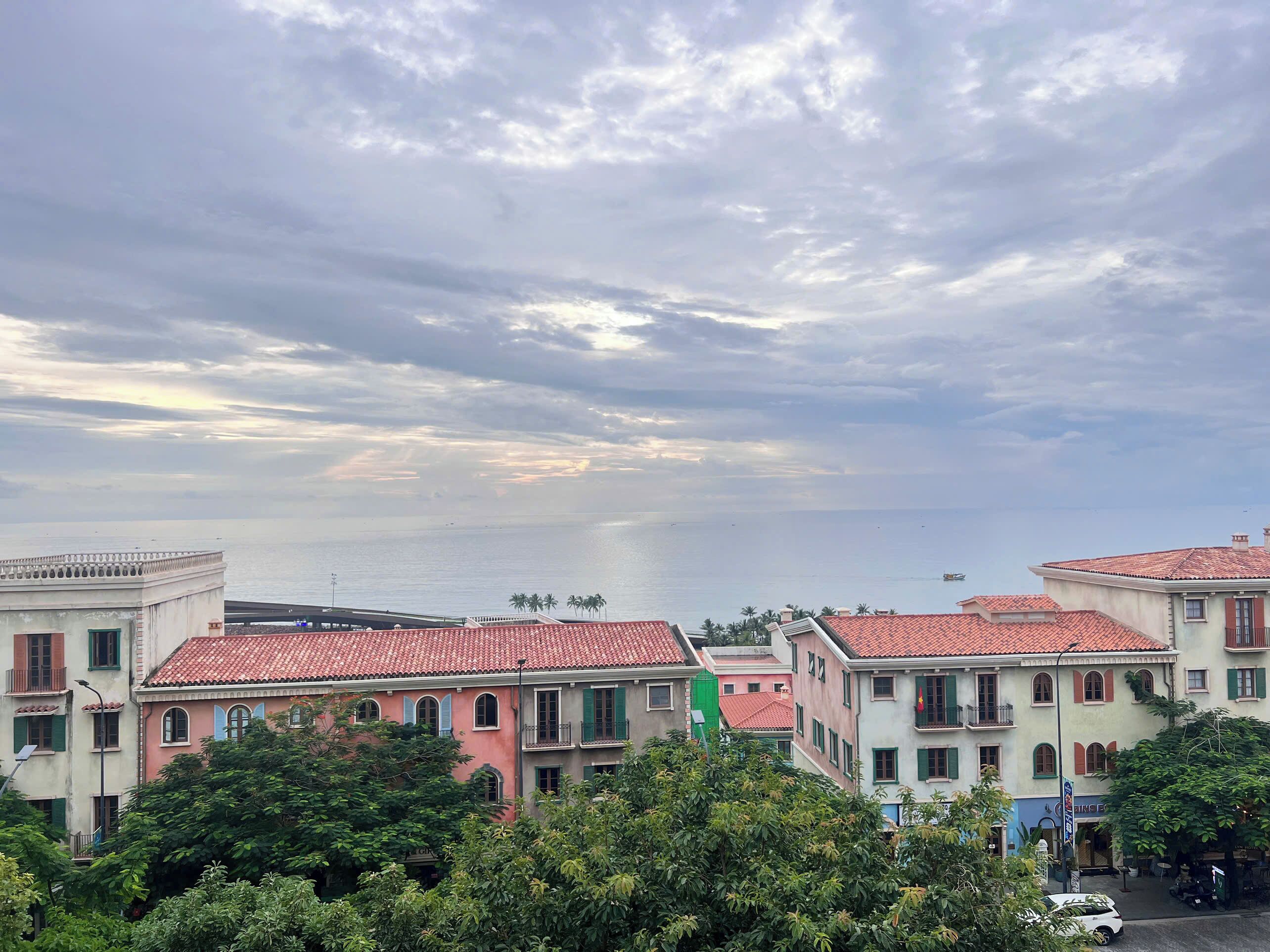 A coastal town with buildings featuring red-tiled roofs and balconies, a clear view of the sea, and lush greenery in the foreground.