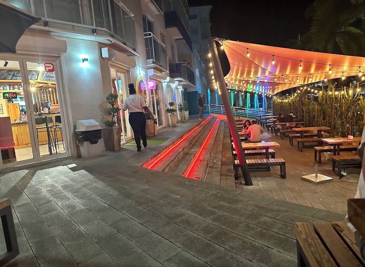 Un patio al aire libre con bancos y mesas de madera, una zona de estar cubierta con luces colgantes y la entrada de un edificio con un letrero de farmacia.