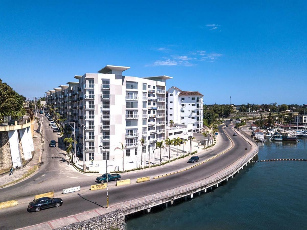 Un immeuble moderne à plusieurs étages, doté de balcons, situé près d’une promenade en bord de mer, avec une route sinueuse et un pont enjambant l’eau.